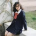Girl in a preppy school uniform with a red bow tie, sitting outdoors, showcasing classic preppy style.