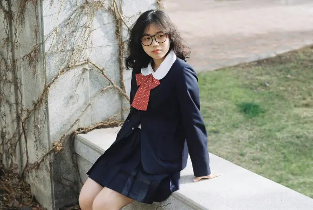 Girl in a preppy school uniform with a red bow tie, sitting outdoors, showcasing classic preppy style.