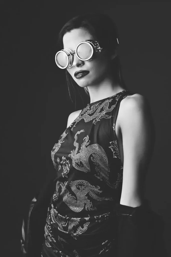 A woman in a black steampunk dress with dragon designs, wearing goggles and dramatic makeup.