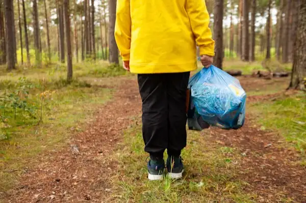 Person in a yellow jacket carrying a trash bag during a forest clean-up.