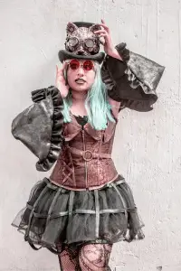 Steampunk woman in a corset and top hat with vibrant blue hair and red goggles.