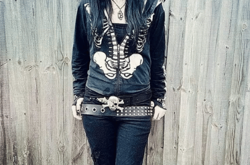 Person wearing emo fashion featuring black skeleton print hoodie, studded belt, dark jeans against weathered wood background